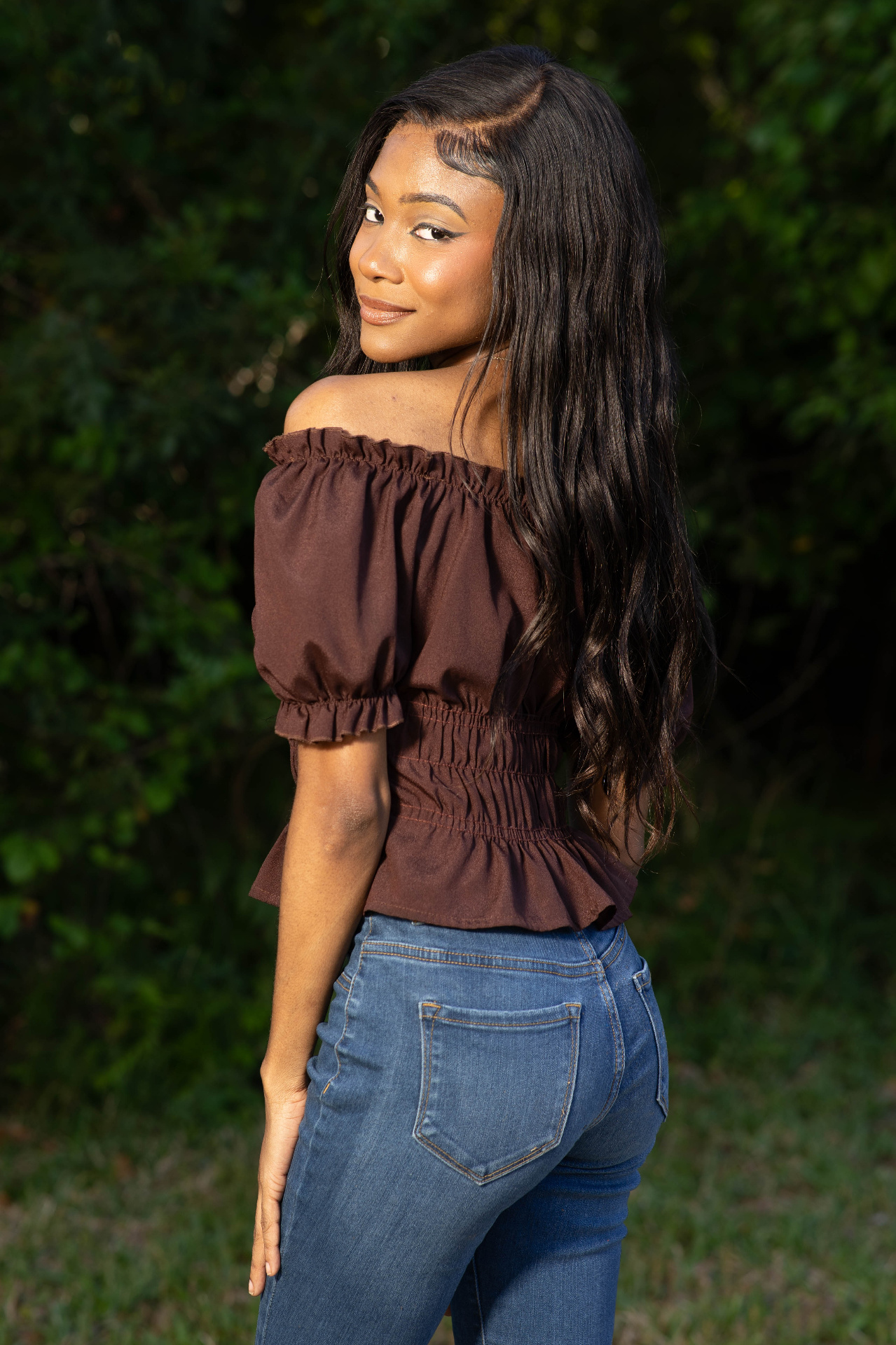 Lifestyle golden hour frame of Chelsey Moné turning over her shoulder in a brown off-shoulder top and denim against greenery