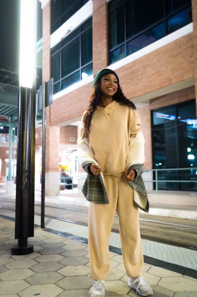 Lifestyle street style frame of Chelsey Moné smiling in a cream sweatsuit and green beanie on a transit platform