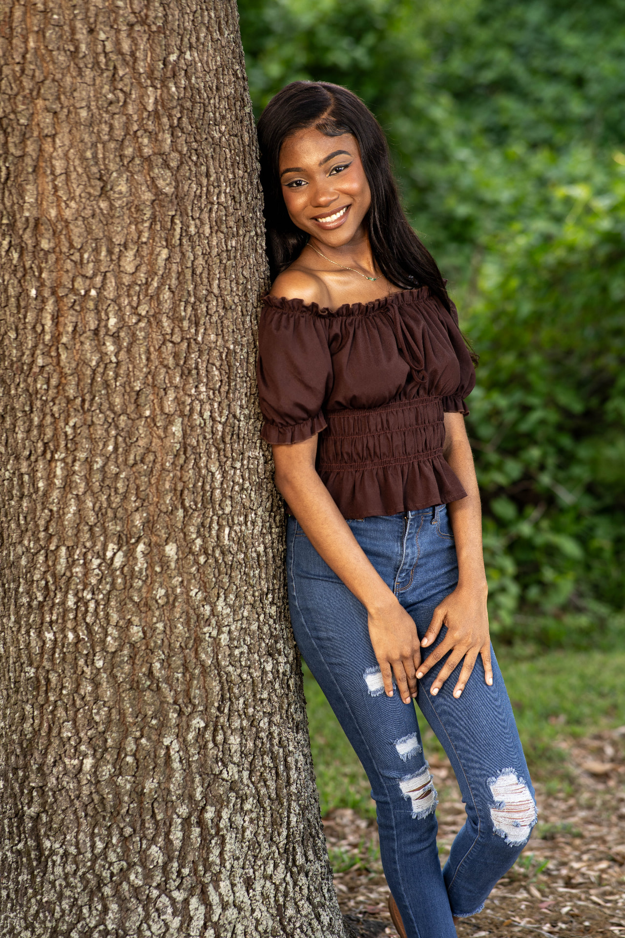 Full-length lifestyle frame of Chelsey Moné leaning against an oak tree in a brown off-shoulder top and denim