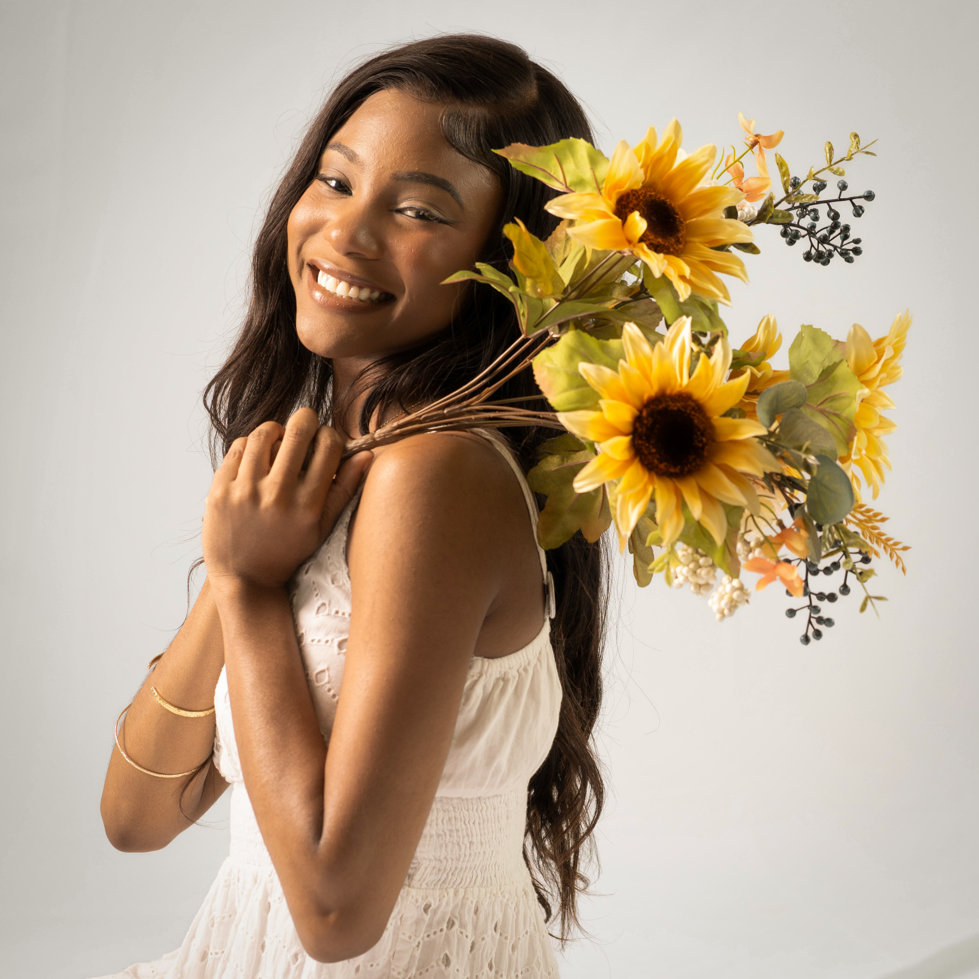 Chelsey Moné smiling in a cream dress holding a bouquet of sunflowers in the studio